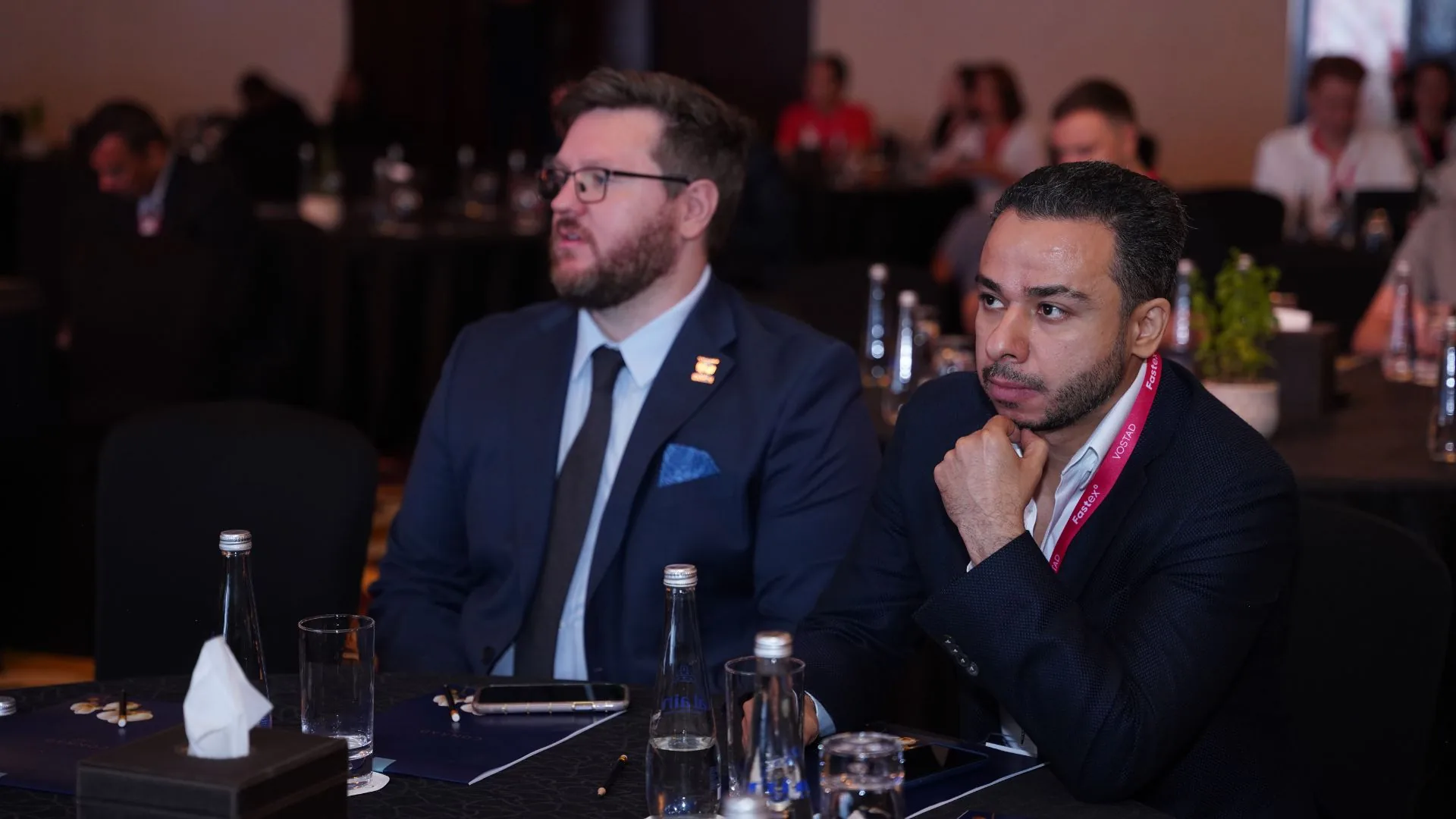 Two delegates listening at a conference table, the room full behind them